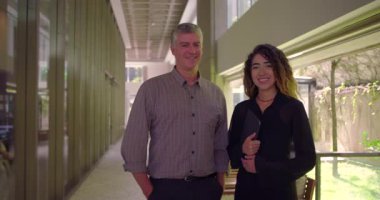 Gülümseyen adam ve kadın ofis koridorunda yan yana duruyor. İkisi de olumlu ifadelerle kameraya bakıyor. İşbirlikçi ve kendine güvenen profesyonel atmosfer.