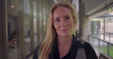 Sakin ve açık bir ifadeyle kameraya bakan, koridorda hareketsiz duran, kendine güveni ve sessizliği temsil eden kadına yakın dur.