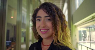 Ofis koridorunda kameraya gülümseyen genç bir kadın, tableti tutarken güvenle duruyor, profesyonel çalışma ortamında rahat duruyor.