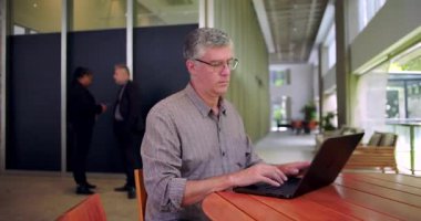 Ortak ofis alanında dizüstü bilgisayarda çalışan, arka planda konuşan, koridorda yuvarlak masada oturan, çağdaş iş ortamında ekrana odaklanan bir adam.