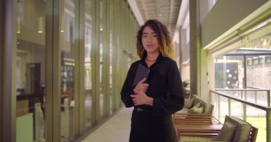 Tablet tutan genç bir kadının tüm vücut portresi modern ofis koridorunda duruyor. Kendine güvenen bir duruşu var. Profesyonel çalışma alanında ileriye bakıyor.