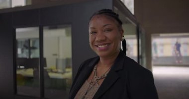 Afro-Amerikalı kadın, cam ofis duvarının yanında durarak samimiyetini, kendine güvenini ve iş ortamındaki yaklaşımını ifade ediyor.