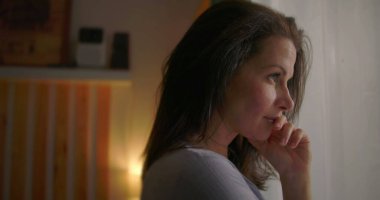 Pencere kenarında duran bir kadın, dudaklara yakın duran, düşünceli bir yüz ifadesi, kapalı alanda tefekkür eden, içe dönük ve sakin bir ruh hali, düşünceli bir an, ev ortamında huzurlu bir yansıma.