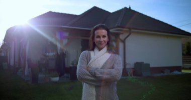 Dışarıda kollarını kavuşturmuş bir kadın, arka bahçede güvenle gülümsüyor, arka planda yeşil bir ev, sıcak atmosferi olan sıradan bir banliyö ortamı.