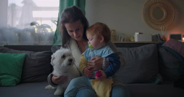 Anne koltukta tüylü beyaz bir köpeği rahatlatırken bebeğini kucağında tutuyor. Bebek emzik ve sıcak bir aile ortamında samimi bir aile anı yaşıyor.