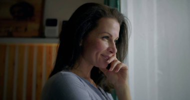 Kadın pencerenin yanında hafifçe gülümsüyor, elleri çenesinde dinleniyor, sakin ve pozitif ruh hali, mutluluk ve düşünceli anlar, kapalı alanda huzur yansıması, hakiki duygu.