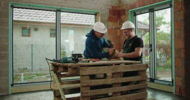 Ahşap iskelet üzerinde işbirliği yapan iki inşaat işçisi, biri matkapla rehberlik ediyor ve ikisi de güvenlik kaskı takıyor, kapalı inşaat alanı inşaatı, inşaat çalışmaları sürüyor.