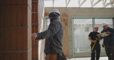 Duvarı kalemle ölçen inşaat işçisi, güvenlik miğferinde odaklanmış miğferle içeride tuğla yapı, hassasiyet ve detaylara dikkat ediyor.