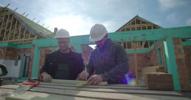 İnşaat alanındaki ahşap kalasları ölçen iki inşaat işçisi, takım çalışması, hassasiyet, güvenlik kaskları, arka planda inşaat halindeki bina