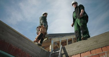 İnşaat işçileri çatıdaki projeyi tartışıyor, bir işçi bina inşaatında yapı, takım çalışması ve iletişimi işaret ediyor, açık hava işbirliği