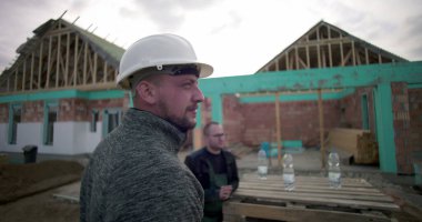 İnşaatta inşaat işçisi, arka planda kısmen inşa edilmiş ev iskeleti, düşünceli bir ifade, açık hava çalışma ortamı.