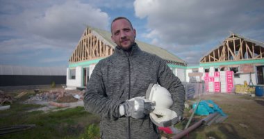 İnşaat işçisi kasketini tutuyor, inşaat sahasında güvenle duruyor, arkaplanında ahşap çerçevesi olan konut inşaatı projesi.