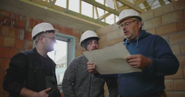 İnşaat halindeki bir binanın planlarını tartışan üç inşaat işçisi, mimari ayrıntılar ve proje planlamasına odaklandı