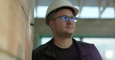 İnşaat işçisi kasketli, düşünceli bir şekilde yan tarafa bakıyor, inşaat alanında kapalı duruyor, duvara yaslanıyor, projeyi düşünüyor.