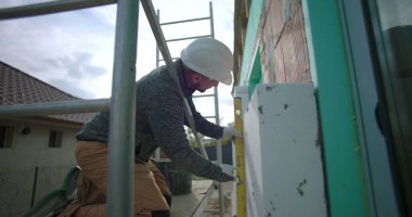 İnşaat işçisi duvardaki yalıtım panelini hizalamak için ruh seviyesini kullanıyor. Hassasiyet, güvenlik kaskı, arka planda yapı iskelesi ve açık hava projesine odaklanmış.