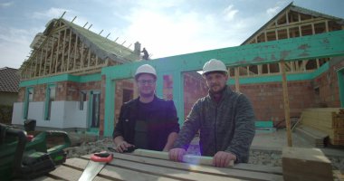 Tamamlanmamış ahşap çerçeve, takım çalışması, kasklar, açık hava inşaat projesinin önünde aletlerle poz veren iki inşaat işçisi.