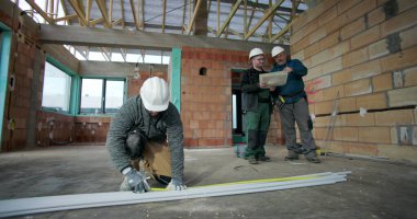 Kattaki PVC borularını ölçen inşaat işçisi, arka plandaki planları, iç inşaat düzenini, takım çalışmasını ve işbirliğini inceleyen iki meslektaşla birlikte