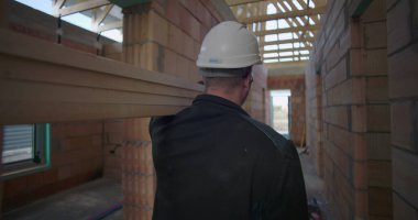 İnşaat işçisi, tamamlanmamış binanın dar koridorunda omuzlarında uzun tahta kiriş taşıyor. İnşaat alanında işçilik ve takım çalışması sergiliyor.