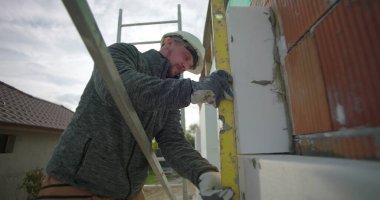 Yalıtım panelini ruh seviyesine ayarlıyorum. İnşaat işçisi iskan duvarına doğru yerleştirme, güvenlik teçhizatı görünür, tuğla zemin.