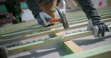 İnşaat işçilerinin çatı kirişlerine çivi çakması, güvenlik eldiveni takması, el ve çekice odaklanması, arka planda meslektaşları olan açık hava inşaat projesi.