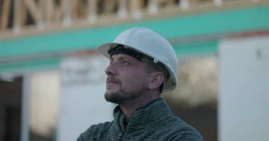 Başlıklı inşaat işçisi, düşünceli bir şekilde şantiyeye bakıyor, bina yapısının geçmişi, odak, planlama ve endüstriyel ithafı temsil ediyor.