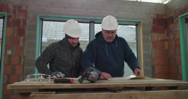 İki inşaat işçisi içerideki ahşap bir masanın planlarını yakından inceliyor, proje detaylarını, güvenlik kasklarını, takım çalışmasını ve iş birliğini tartışıyorlar.