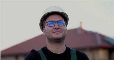 Başlık ve gözlük takan inşaat işçisi, yan profil görüntüsü, iş yerinde gülen, arka planda açık çerçeve yapısı, uzmanlık ve memnuniyeti temsil eden.