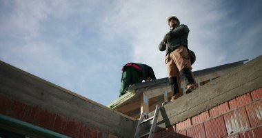 İnşaat işçileri merdivende çatı yapısını güvence altına alıyorlar, inşaat sahasına, güvenlik ekipmanlarına, iskeleye, mavi gökyüzüne, güneşli bir günde endüstri çalışanlarına odaklanmış durumdalar.