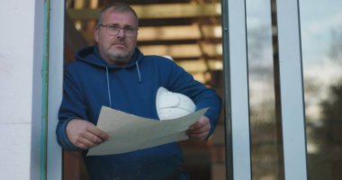 İnşaat işçisi elinde odaklı bir ifadeyle proje planlarını inceliyor, kapı önünde duruyor, inşaat işlerinde hassasiyete adanmış.