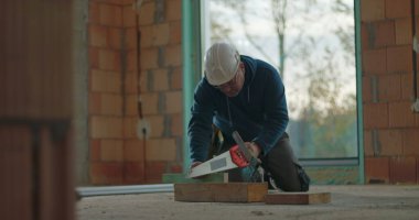 İnşaat işçisi ev inşaatında el testeresiyle odun kesiyor ve diz çöküyor. İşine odaklanıyor. Etrafı tamamlanmamış duvarlarla çevrili.