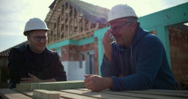İnşaat alanında sohbet eden iki inşaat işçisi proje detayları, konut inşaatında takım çalışması ve işbirliği, arka planda ev iskeleti tartışmaları yapıyorlar.