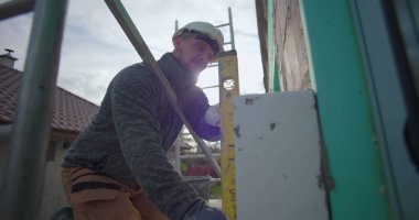 Dış duvardaki yalıtım panelini ruh seviyesine ayarlıyorum. İnşaat işçisi ev işleriyle uğraşıyor. Tuğla ve yalıtım düzenini kapatıyor.