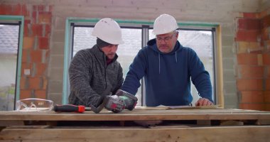 İki inşaat işçisi ahşap bir masanın üzerindeki bina planlarını gözden geçiriyorlar, içerideki yenileme projesini tartışıyorlar, yetenekli işçiler arasındaki işbirliği, güvenlik kaskları, takım çalışması