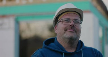 Sert şapkalı inşaat işçisi düşünceli bir ifadeyle bir inşaat projesinde, açık havada bir yere bakıyor.