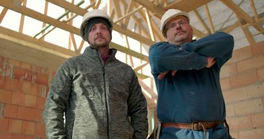 Sert şapkalı iki inşaat işçisinin, takım çalışması, birlik ve profesyonelliği temsil eden kısmen inşa edilmiş bir yapının içinde güvenle durduğu alçak açılı bir bakış açısı.