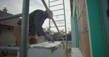 İnşaat işçisi, yalıtımlı paneli yapıştırıcıyla kaldırıyor. İnşaat alanındaki duvar döşemesi, yapı iskelesi ve arka plandaki tuğla duvar için hazırlanıyor.