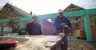 Ahşap blokta testereyle çalışan inşaat işçisi, meslektaş gözlemleme, inşaat alanında takım çalışması, arka planda ev iskeleti, hassasiyet ve güvenliğe odaklanma