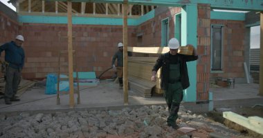 İnşaat alanında omuzlarında ahşap kalaslar taşıyan işçiler, takım çalışması ve marangozluk görevlerine odaklanmış durumda, görünür yapı ve kirişlerle yerleşim alanı inşaatı sürüyor