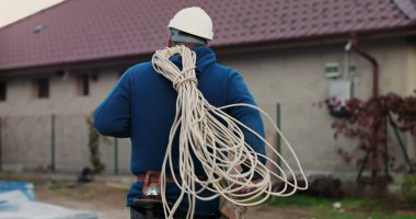 İnşaat alanında omuz üzerinde paketlenmiş elektrik kabloları taşırken, miğferli işçi araçları yönetirken ve kurulum için hazırlanırken binaya doğru yürüyordu.