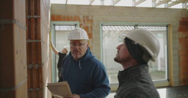 İnşaat işçisine açıklarken tuğla duvarı işaret eden mühendis, arka planda açık yapı ve geniş pencerelerle ilgili ayrıntıları tartışıyor