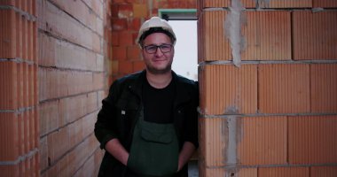 İnşaat işçisi bitmemiş binanın içinde güvenle duruyor, tuğladan duvara yaslanıyor, miğfer ve gözlük takıyor, rahat poz veriyor, yapım aşamasında.