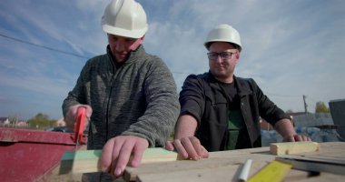 İki inşaatçı birlikte çalışıyor, biri odun kesmek için el testeresi kullanırken diğer saatler, etkin inşaat sahasında takım çalışması, yapı iskeleti ve aletler etrafında.