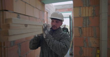 İnşaat işçisi, tamamlanmamış iskan binasının koridorundaki ahşap kalasları kaldırıyor. İşgücü ve inşaat alanında hassasiyet odaklanıyor.