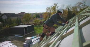 Konut inşaatı sırasında çatıya çivi çakan işçiler, güvenlik ve hassasiyete odaklanmış, başlık ve eldiven takıyor, marangozluk ve çatı projesi sürüyor