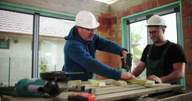 Ahşabı delen iki inşaat işçisi, şantiyedeki iskeleti güvence altına almak için işbirliği, hassasiyet ve doğruluk odaklı, her ikisi tarafından takılan güvenlik kaskları