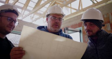 Üç inşaat işçisi gülümseyip bina planlarını tartışırken, inşaat projesiyle ilgili kısmen inşa edilmiş bir yapının planlarını gözden geçiriyorlar.