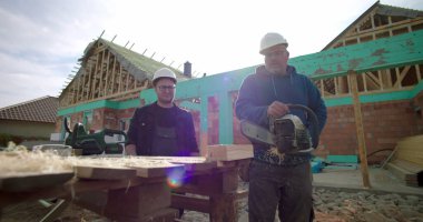 Ahşap blokta testereyle çalışan inşaat işçisi, meslektaş gözlemleme, inşaat alanında takım çalışması, arka planda ev iskeleti, hassasiyet ve güvenliğe odaklanma