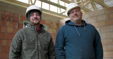 İnşaat alanında poz veren iki inşaat işçisi, güvenlik kaskı takan adamlar tamamlanmamış yapıda yan yana duruyorlar, takım arkadaşlığı ve iş gururu.