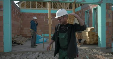 İnşaat alanında omuzlarında ahşap kalaslar taşıyan işçiler, takım çalışması ve marangozluk görevlerine odaklanmış durumda, görünür yapı ve kirişlerle yerleşim alanı inşaatı sürüyor