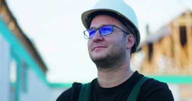 Başlık ve gözlük takan inşaat işçisi, yan profil görüntüsü, iş yerinde gülen, arka planda açık çerçeve yapısı, uzmanlık ve memnuniyeti temsil eden.